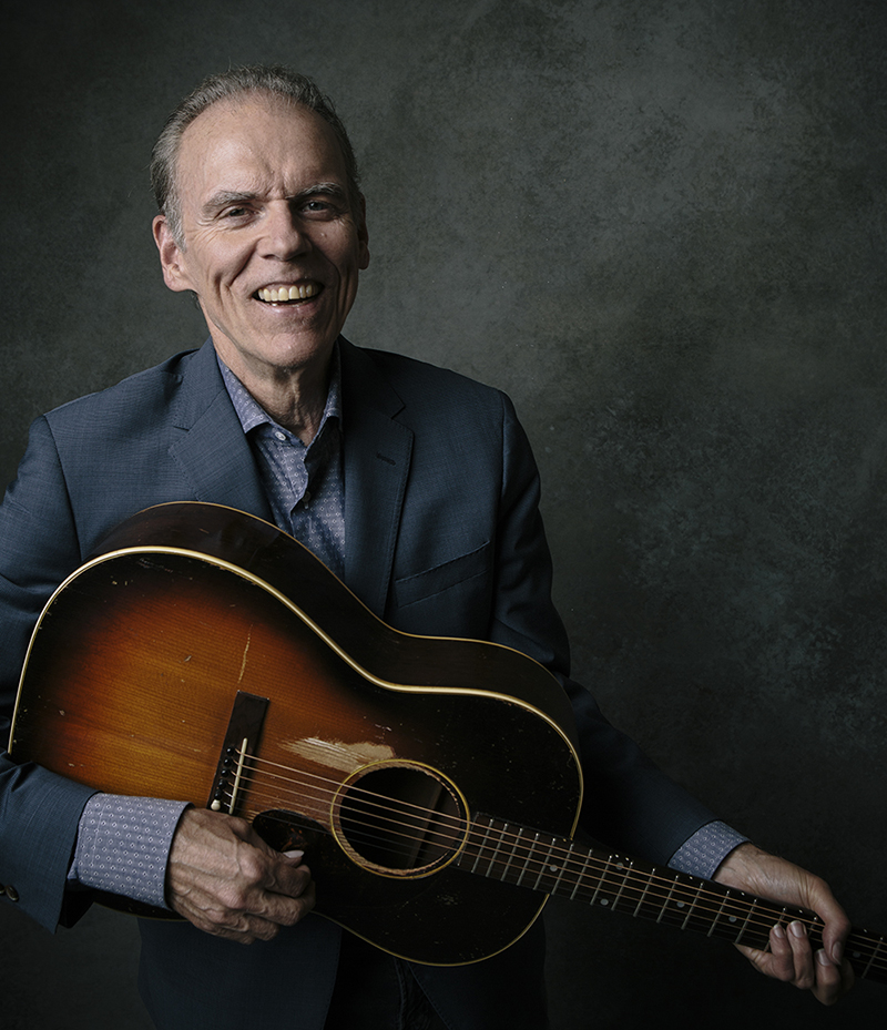 John Hiatt, Man in blue blazer posing with weathered acoustic guitar in front of grey/green marbled backdrop